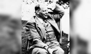 Ein schwarzweiß Bild von einem Mann, er trägt einen Anzug und hebt die linke Hand zum Kopf