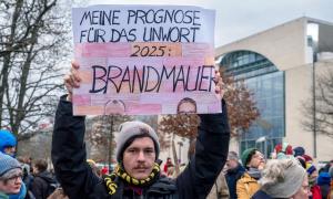 Ein Mann mit Mütze hält ein Schild hoch, auf dem steht: "Meine Prognose für das Unwort 2025: Brandmauer". Auch die jeweilige Stirn von Friedrich Merz und Alice Weidel ist zu sehen.