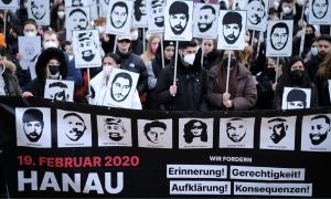 Menschen vor einem Transparent, auf dem acht Gesichter zu sehen sind, sie halten Schilder hoch, auf denen auch Gesichter zu sehen sind