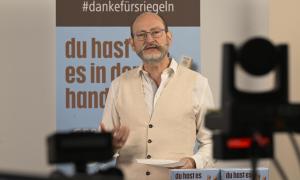 Ein Mann mit Brille steht vor Kameras, hinter ihm ist ein Plakat mit Schokoriegeln zu sehen.