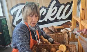 ältere Frau mit halblangen grauen Haaren und orangener Schürze gibt Brötchen aus