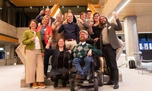 12 Menschen stehen oder sitzen vor einer Treppe, sie halten Gläser hoch