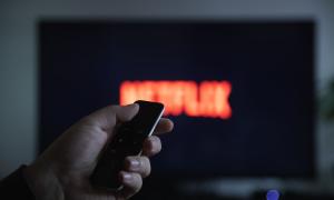 Es ist im Hintergrund ein Fernseher zu sehen, der unfokussiert das Netflix-Logo zeigt. Im Vordergrund ist eine Hand, die eine Fernbedienung hält.