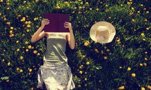 Eine Frau liegt auf einer Wiese, ein Buch vor dem Gesicht, neben ihr liegt ein Strohhut