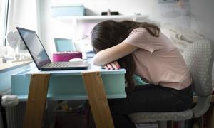 Ein Mädchen sitzt am Tisch und hat den Kopf in den Armen vergraben, vor ihr ein aufgeklapptes Laptop