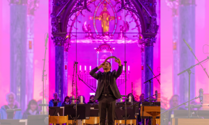 Ein Mann in schwarzem Anzug bläst in einem farbig beleuchteten Kirchenraum in ein gebogenes Horn. Im Hintergrund sind Notenständer und sitzende Menschen zu sehen.