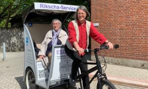Eine Rikscha mit der Aufschrift "Rikscha-Service" und einer älteren Frau, die als Fahrgast darin sitzt und einer Frau, die als Fahrerin vorne auf dem Fahrrad sitzt