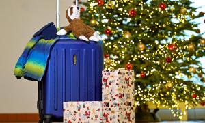 Kinderjacke, Handschuhe und Stofftier liegen auf einem gepackten Rollkoffer, vor dem Geschenke auf dem Boden stehen, im Hintergrund ein geschmückter Weihnachtsbaum