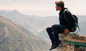 Ein Mann mit Rucksack sitzt an einer Kante, im Hintergrund Berge